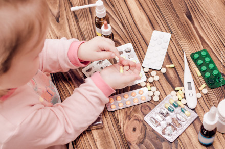 Kleines Kind im rosa Schlafanzug sitzt an einem Holztisch & hält eine gelbe Kapsel in der Hand. Auf dem Tisch liegen verschiedene Medikamente, darunter Tabletten in Blisterpackungen, Kapseln, ein digitales Fieberthermometer & medizinische Tropfflaschen.