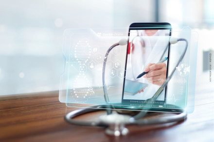 Ein Stethoskop liegt auf einem Holztisch, während im Hintergrund ein Tablet oder Smartphone zu sehen ist, das einen Arzt zeigt, der auf einem digitalen Gerät notiert.
