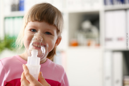 Ein kleines Mädchen in einem rosa Oberteil verwendet einen Inhalator mit einer Gesichtsmaske. Sie lächelt in die Kamera und befindet sich in einem gut beleuchteten Raum mit Regalen im Hintergrund.