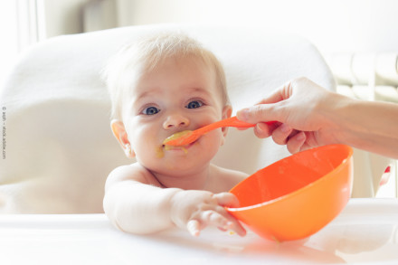 Ein Baby sitzt in einem Hochstuhl und wird mit einem orangefarbenen Löffel aus einer orangefarbenen Schüssel gefüttert. Das Baby hat ein wenig Essen um den Mund herum. Eine Hand hält den Löffel, während das Baby zuschaut und die Schüssel festhält.