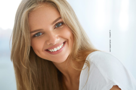 Eine junge Frau mit langen blonden Haaren und blauen Augen lächelt in die Kamera. Sie trägt ein weißes Oberteil. Der Hintergrund ist verschwommen und in hellen Farben gehalten.