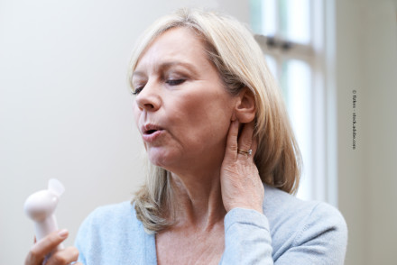 Eine ältere Frau hält sich den Nacken und benutzt einen kleinen Handventilator, um sich abzukühlen. Sie trägt einen hellblauen Pullover und hat blonde Haare. Sie steht in einem hellen Raum.