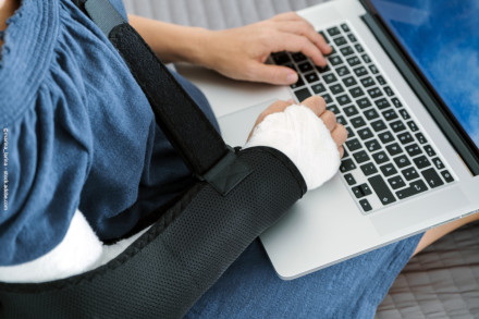 Eine Person mit einem blauen Oberteil tippt auf einer Laptop-Tastatur. Ihr rechter Arm ist in einem Verband und einer Armschlinge fixiert.