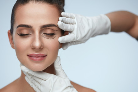 Eine Frau mit geschlossenen Augen. Sie hat markierte Linien unter einem Auge und wird von Händen in weißen medizinischen Handschuhen sanft am Kinn und an den Schläfen gehalten, was auf eine kosmetische Behandlung hinweist.