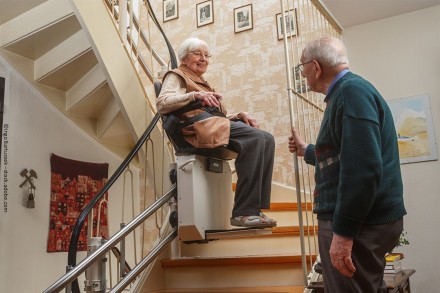 Ein älteres Paar in einem Haus. Eine Frau sitzt auf einem Treppenlift und fährt die Treppe hinauf, während ein Mann neben dem Treppenlift unten an der Treppe steht. Die Frau lächelt und schaut den Mann an.