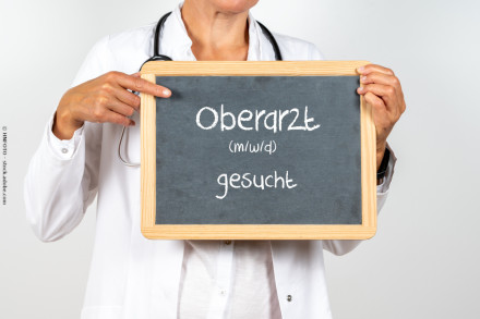 Eine Person in einem weißen Arztkittel hält ein kleines Tafelschild. Auf der Tafel steht in weißer Kreideschrift: "Oberarzt (m/w/d) gesucht". Die Person zeigt mit einem Finger auf das Schild.