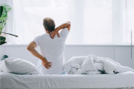 Ein Mann sitzt auf einem Bett. Er hält seinen Rücken & seine Hüfte, als ob er Schmerzen hätte. Das Licht fällt durch große Fenster mit weißen Vorhängen in den Raum. Neben dem Bett befindet sich eine Pflanze. Das Bett ist unordentlich mit Kissen & Decken.