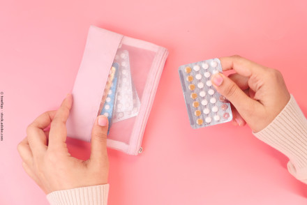 Eine Person hält eine Blisterpackung mit Tabletten in der einen Hand und greift mit der anderen Hand nach einer rosa Tasche, die ebenfalls Tablettenpackungen enthält. Der Hintergrund ist rosa.