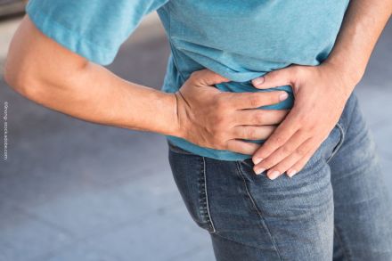 Ein Mensch trägt ein hellblaues T-Shirt und dunkelblaue Jeans und hält sich mit beiden Händen den rechten unteren Bauchbereich, als ob er Schmerzen hat. Der Hintergrund ist unscharf. Dieses Bild vermittelt den Eindruck von Bauchschmerzen oder Unwohlsein.