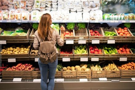Eine Frau mit braunem Rucksack steht vor einem Gemüseregal in einem Supermarkt. Das Regal ist mit verschiedenen frischen Gemüsesorten wie Kartoffeln, Tomaten, Paprika und Karotten gefüllt.
