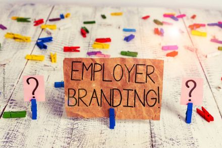 Ein handgeschriebenes Schild mit der Aufschrift "EMPLOYER BRANDING!" steht auf einem hellen Holztisch. Links und rechts des Schildes befinden sich zwei rosa Karten mit einem Fragezeichen darauf, gehalten von blauen Wäscheklammern.
