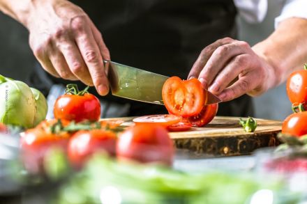 Ein Mann schneidet eine Tomate mit einem scharfen Küchenmesser auf einem Holzbrett. Im Vordergrund und Hintergrund sind weitere Tomaten und grünes Gemüse zu sehen.