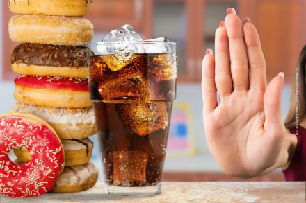 Eine Nahaufnahme von einem Stapel Donuts mit verschiedenen Glasuren und einem Glas Cola mit Eiswürfeln. Neben dem Glas ist eine Hand zu sehen, die eine "Stop"-Geste macht.