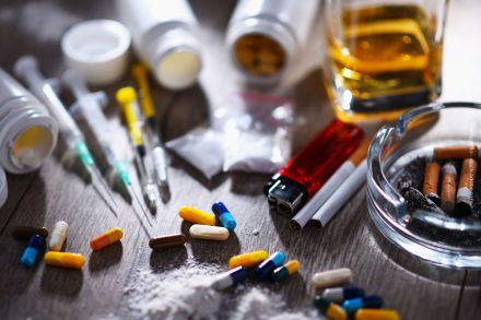 Uunordentlicher Tisch mit verschiedenen Drogen und Drogenutensilien. Es gibt Pillen, Spritzen, Tablettenfläschchen, ein Feuerzeug, Zigarettenstummel in einem Aschenbecher und ein Glas. Auch kleine Plastiktüten und Pulver sind zu sehen.