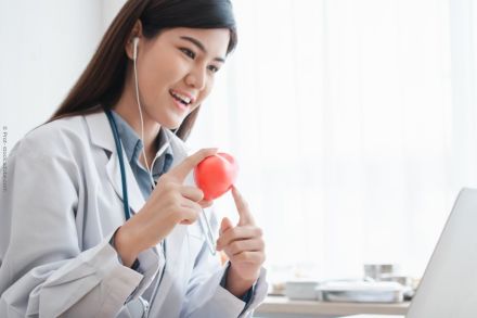 Eine Ärztin sitzt an einem Schreibtisch und trägt Kopfhörer, während sie mit einem Patienten über einen Laptop spricht. Sie hält ein rotes Modell eines Herzens in der Hand und erklärt möglicherweise eine medizinische Angelegenheit.
