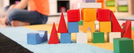 Bauklötze bilden einen Turm in einem Spielzimmer. Im Hintergrund sitzt ein Kind im Schneidersitz und schaut nach links.