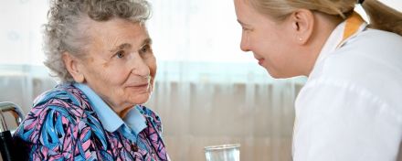 Pflegekraft spricht liebevoll mit einer älteren Frau im Rollstuhl und reicht ihr ein Glas Wasser.