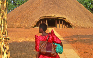 Frau in bunter Kleidung steht vor reetgedecktem Haus in Kampala, Uganda, Afrika