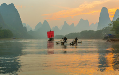 Fisherman stands on traditional bamboo boats at sunrise (boat with a red sail in the background)