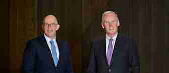 Two men in suits stand smiling against a dark wooden background. The man on the left wears glasses and a blue tie, while the man on the right wears a purple tie. Both have their hands in their pants pockets.