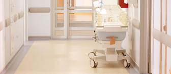 A hospital hallway with a neonatal incubator on wheels positioned near a closed door, under bright overhead lighting, and surrounded by white walls and medical equipment.