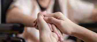 A close-up of two people holding hands. One person, slightly blurred in the background, is sitting in a wheelchair, and the other person, visible only by their hands, is providing support and comfort. The mood appears to be compassionate and supportive.
