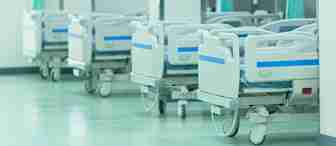 A row of empty hospital beds with white sheets and blue accents are lined up against a wall in a brightly lit room. Green privacy curtains are partially drawn between each bed, and the floor is clean and shiny. The atmosphere appears sterile and well-maintained.