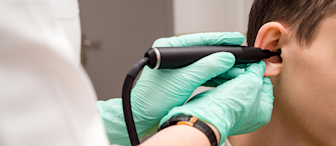 A healthcare professional wearing green gloves uses an otoscope to examine the ear of a young person in a yellow shirt. The focus is on the professional's hand and the otoscope, with the patient's ear and part of their head visible.