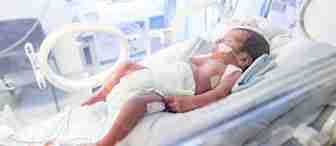 A newborn baby lies in an incubator in a neonatal intensive care unit (NICU). The baby has monitoring sensors attached to their body and is wearing a diaper. Medical equipment and tubes surround the incubator, indicating the baby's need for specialized care.