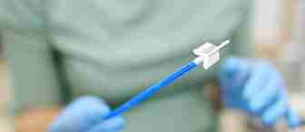 A person wearing blue gloves holds a cervical screening brush, commonly used for Pap smear tests, with both hands. The background is blurred.