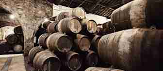 A dimly lit cellar with stacks of large wooden barrels, some showing signs of age. The stone walls and archways add a rustic feel to the space. The barrels are positioned haphazardly, contributing to the overall vintage atmosphere of the old cellar.