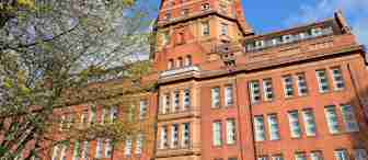A large red-brick building with multiple windows and a distinctive central tower stands against a bright blue sky. Trees with budding leaves frame the left side of the image. The architecture is classic and intricate, indicating an older, possibly institutional building.