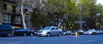 A busy city street with cars driving past. The foreground features blurred pedestrian crossing lines. Trees and buildings line the street, and the background shows more parked cars and city architecture illuminated by evening lights.