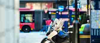 Parked motorcycles on a city street with a red bus and pedestrians in the background. The scene includes street signs and a map on the right, with bollards lining the sidewalk.