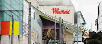 People walking up steps towards a large shopping center entrance with the "Westfield" sign above. The building features colorful panels and large advertisements. There are trees and greenery on the right side.
