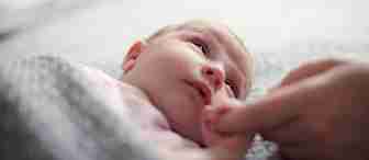 A baby lies on a soft, grey blanket, looking up. The baby is wrapped up in the blanket, and a person’s hand is gently holding the baby’s hand. The scene evokes a sense of warmth and tenderness.