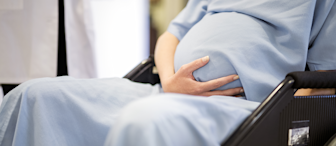 A pregnant person in a light blue hospital gown sits in a wheelchair, gently cradling their baby bump with one hand. A healthcare professional, partially visible and dressed in white, stands nearby. The background is softly blurred.
