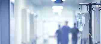 A close-up of an intravenous (IV) drip in focus, hanging on a stand in a hospital corridor. The background is blurred, showing medical staff in scrubs walking down the brightly lit hallway.