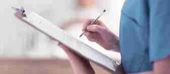 A healthcare worker in blue scrubs is holding a clipboard and writing with a pen. The clipboard contains some documents, and the background appears to be a blurred hospital or medical setting. The focus is on the worker’s hands and the clipboard.