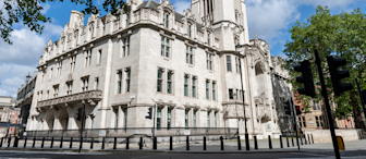A picture of the UK Supreme Court building in London on a sunny day, featuring its ornate stone facade and tower. The foreground includes traffic lights, a few trees, and a section of pavement. The sky is partly cloudy with patches of blue.