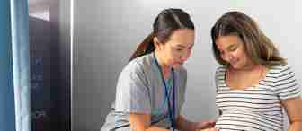 A healthcare professional in gray scrubs examines a pregnant woman in a striped dress, gently placing hands on her belly in a medical setting. The room includes an examination table and blue privacy curtains.