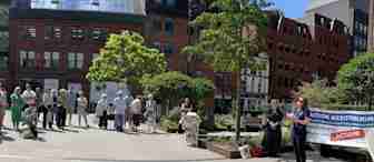 A group of people stands in a sunny plaza, attentively listening to a woman speaking at a microphone. A large banner reading "ACTION MESOTHELIOMA" is displayed to the right. Surrounding buildings and a few trees are visible in the background.
