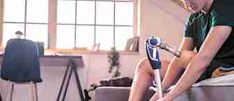 A person with a prosthetic leg sits on a couch in a well-lit room, adjusting the prosthetic. They are wearing casual clothes and sneakers. In the background, there's a desk, a chair, and a rocking horse. The room has a friendly and cozy ambiance.