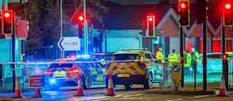 A nighttime scene shows a road closure with traffic lights on red and police vehicles with flashing lights blocking the street. Cones and barriers are set up, and several emergency personnel in reflective uniforms are gathered by a building in the background.