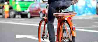 A person wearing jeans and black shoes is riding an orange bicycle on a city street. The street is marked with directional arrows and a "BUS" lane sign. Vehicles and a mural are visible in the background.