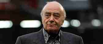 An elderly man with a receding hairline is wearing a dark suit jacket over a patterned shirt. He is outdoors, with blurred lights in the background.