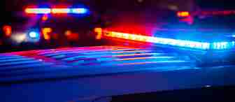 Close-up of a police car with a flashing red and blue light bar at night. Blurred lights from other emergency vehicles are visible in the background, casting a mix of blue and red hues across the scene. The atmosphere appears tense and urgent.