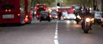 Urban street scene with blurry red double-decker buses, cars, and a motorcyclist with headlights on riding towards the camera. Trees line the street in the background, and the road markings lead the eye towards the horizon.