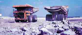 Two large heavy-duty trucks positioned on a rocky construction or mining site. The trucks, facing away from each other, show dusty tires and an open bed, surrounded by a landscape of rocks under a blue sky with scattered clouds.