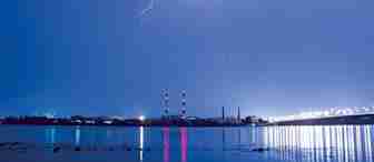 A nighttime scene shows lightning illuminating a cloudy sky over an industrial area. Two tall chimneys with red and white stripes stand prominently. Their reflections, along with numerous lights from the area, shimmer on the calm water below.